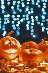 Pumpkin with smiling face placed next to fresh pumpkins on the table with leaves and candles in the real photo with blurred background