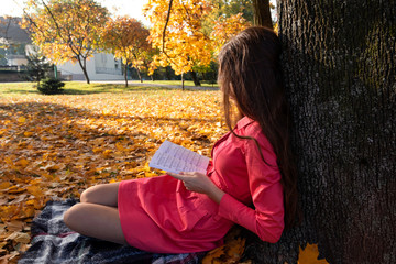 Beautiful young brunette girl sitting on fallen autumn leaves in park, reading book.