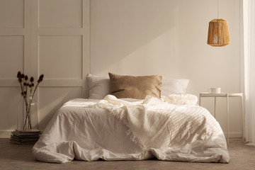 Fototapeta na wymiar Pillows on white bed between flowers and table in minimal bedroom interior with lamp. Real photo