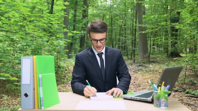 Inspired businessman writing his ideas in notebook, sitting at desk in forest