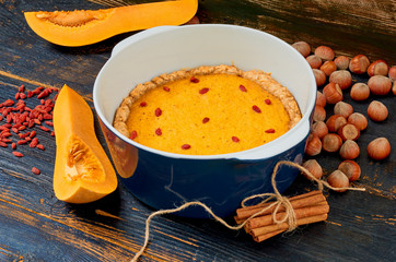 Thanksgiving pie with goji berry in the baking dish decorated with fresh pumpkin slices, cinnamon sticks and hazelnuts on the dark wooden rustic background. Side view 