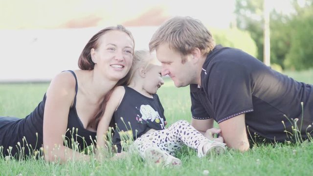 Dad, mom and little daughter are sitting on the green grass in the park. Parents kiss the baby and play with her.