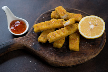 Fried fish sticks or breaded fish fillet with lemon and sauce on a rustic wooden serving board, horizontal shot
