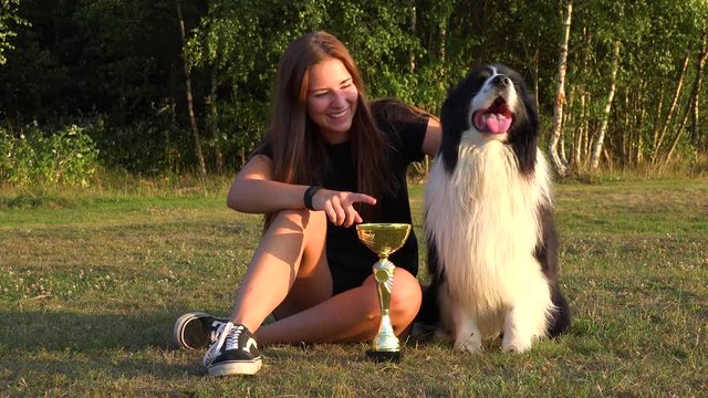 A young woman and a Border Collie sit in a meadow with a trophy, the woman shows the trophy to the dog and laughs