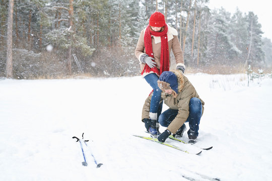 Full length portrait of young couple enjoying ski walk in winter forest during date, focus on man helping girlfriend put on her skis, copy space