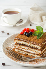 Delicious Tiramisu cake with fresh berries and mint on a plate on a light background. Breakfast with a Cup of coffee.