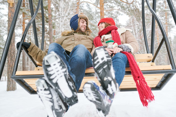 Full length low angle portrait of happy young couple sitting on wooden swing and looking at each other tenderly while enjoying date in winter forest