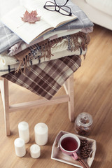 Fototapeta na wymiar Plaids. A cup of hot tea. Autumn cozy interior. On a wooden chair is a stack of warm blankets. Candles, leaves, cones, basket, cinnamon. Book and glasses. Autumn. Winter.