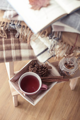 Plaids. A cup of hot tea. Autumn cozy interior. On a wooden chair is a stack of warm blankets. Candles, leaves, cones, basket, cinnamon. Book and glasses. Autumn. Winter.