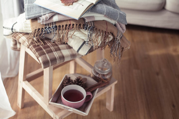 Plaids. A cup of hot tea. Autumn cozy interior. On a wooden chair is a stack of warm blankets. Candles, leaves, cones, basket, cinnamon. Book and glasses. Autumn. Winter.