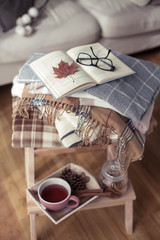 Fototapeta na wymiar Plaids. A cup of hot tea. Autumn cozy interior. On a wooden chair is a stack of warm blankets. Candles, leaves, cones, basket, cinnamon. Book and glasses. Autumn. Winter.