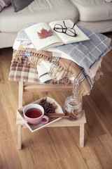 Obraz na płótnie Canvas Plaids. A cup of hot tea. Autumn cozy interior. On a wooden chair is a stack of warm blankets. Candles, leaves, cones, basket, cinnamon. Book and glasses. Autumn. Winter.