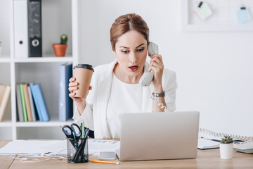 beautiful shocked businesswoman holding coffee to go while talking on phone and working with documents and laptop at workplace