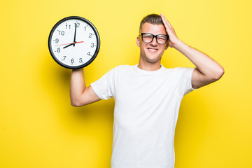 Portrait of young man holding a clock with his hands on head forget something over yellow background