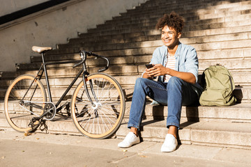 Cheerful young african guy sitting outdoors with bicycle on the street on steps using phone.