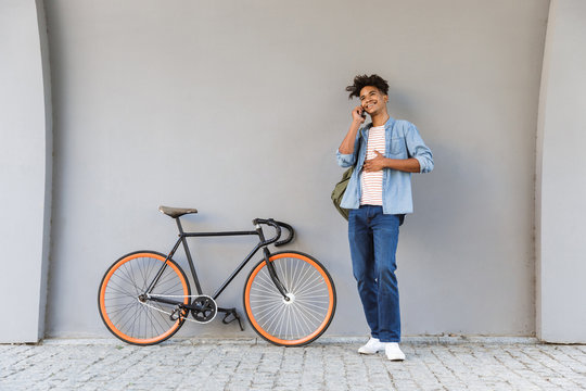 Happy young african guy walking outdoors with bicycle talking by mobile phone.