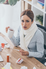Fototapeta na wymiar high angle view of sick businesswoman in scarf holding cup of tea and electric thermometer sitting at office and looking at camera