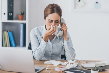 close-up portrait of ill adult businesswoman with runny nose talking by wired phone at office