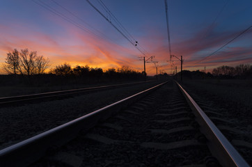 Obraz na płótnie Canvas Dark silhouettes railway infrastructure against background of dramatic sunset. View of railroad going straight away to sun and beyond the horizon. The colorful sky with clouds at sundown