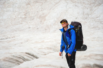 Traveler in a cap and sunglasses with a backpack on his shoulders in the snowy mountains on the glacier against the sky and clouds. Traveler in a natural environment