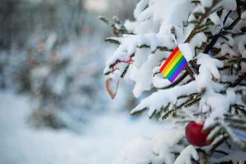 LGBT flag. Christmas background outdoor. Christmas tree covered with snow and decorations and rainbow flag.  New Year / Christmas holiday greeting card. - 228094315
