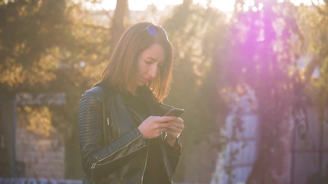 Attractive young girl stands in middle of busy buzzling city on summer sunset. Gorgeous young woman in a bright sunlight uses her phone.