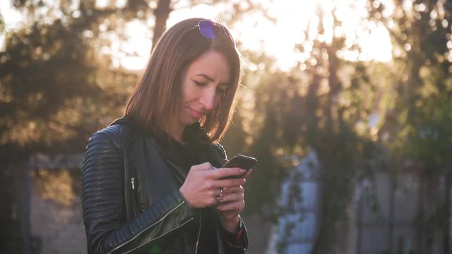 Attractive young girl stands in middle of busy buzzling city on summer sunset. Gorgeous young woman in a bright sunlight uses her phone.