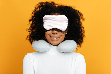 Comfort sleep. Woman portrait. Afro American girl in sleep mask and with a neck pillow is smiling, on a yellow background