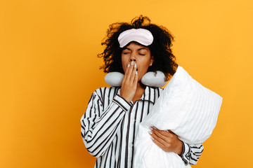 Sleeping. Dreams. Woman portrait. Afro American girl in pajama and sleep mask is holding a pillow and yawning, on a yellow background - obrazy, fototapety, plakaty
