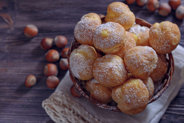 Fresh handmade profiteroles in a wicker basket on a lace napkin and several hazelnuts are scattered alongside on a wooden table