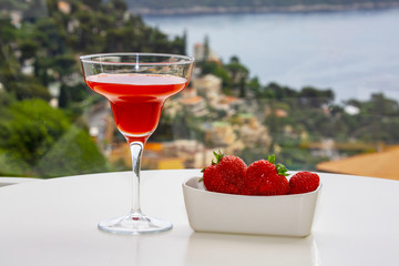 A glass with a red drink and strawberries against the backdrop of the mountains and the sea.