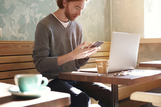 Handsome businessman with long hair wearing sweater calling by smartphone sitting in sunny cafe, using laptop having coffee alone.