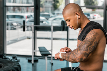 side view of young shirtless sportsman in earphones using smartwatch in gym