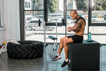 muscular shirtless african american sportsman in earphones sitting and using smartwatch in gym