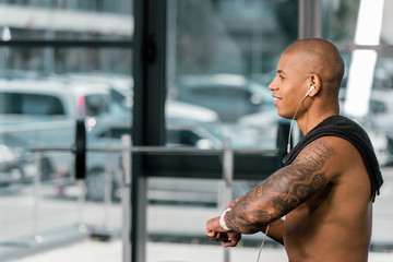 side view of smiling shirtless sportsman in earphones using smartwatch in gym