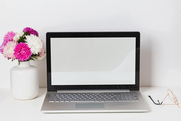 Fototapeta premium Feminine workplace with laptop with white empty screen and flowers in the vase