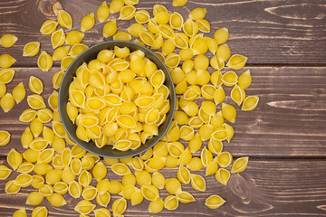 Lot of whole raw yellow pasta conchiglie variety in a grey ceramic bowl flatlay on brown wood