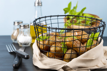 Baked potatoes in a black basket. Knife and fork. Basil, olive oil, salt, pepper. The background is light blue. Copy space. Horizontal shot.