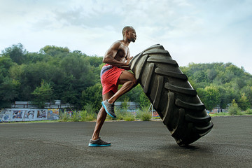 Handsome african american muscular man flipping big tire outdoor.