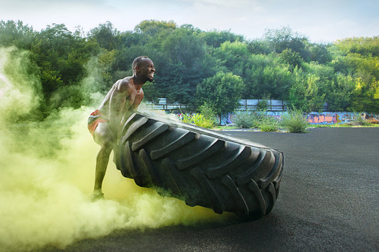 Handsome african american muscular man flipping burning big tire outdoor with smoke - Powered by Adobe