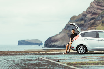Young handsome man sitting in car trunk and enjoy beauty view of rocks and ocean. Car trip. Summer travel. © F8  \ Suport Ukraine