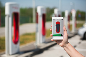 Hand with phone on a background of electric car charging station. On a divice screen indicator of...