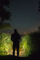 A man with a flashlight looking at the stars above the lake and a meteor falling in the distance