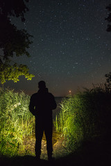 A man with a flashlight looking at the stars above the lake and a meteor falling in the distance