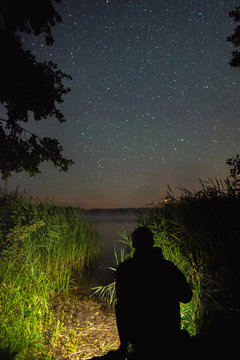 A man with a flashlight looking at the stars above the lake and a meteor falling in the distance