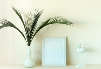 mock up of white photo frame on pale beige  background, tropical palm leaves in a vase and interior decor bird. Copy space
