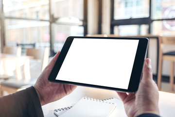 Mockup image of hands holding and using black tablet pc with blank white desktop screen with notebooks on the table