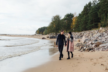Young couple in love walking on the beach coast. Cold autumn weather, trees in the background. Young man wearing a wool sweater, girl in a pink coat