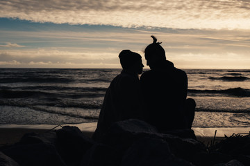 Happy romantic couple sitting on the beach near ocean, relaxing, holding around each other. Silhouettes, sunset, back view