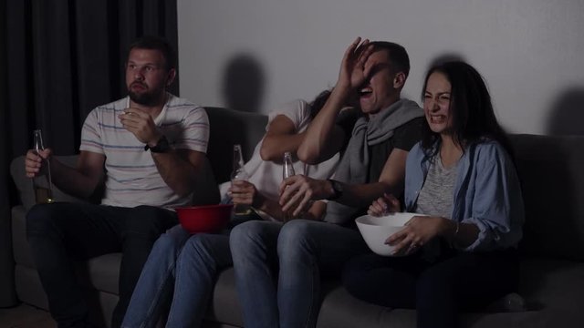 Four friends eating popcorn, drinking beer watch horror movie together and are very captivated and scared, sit still. Close eyes from scary picture. Movie night